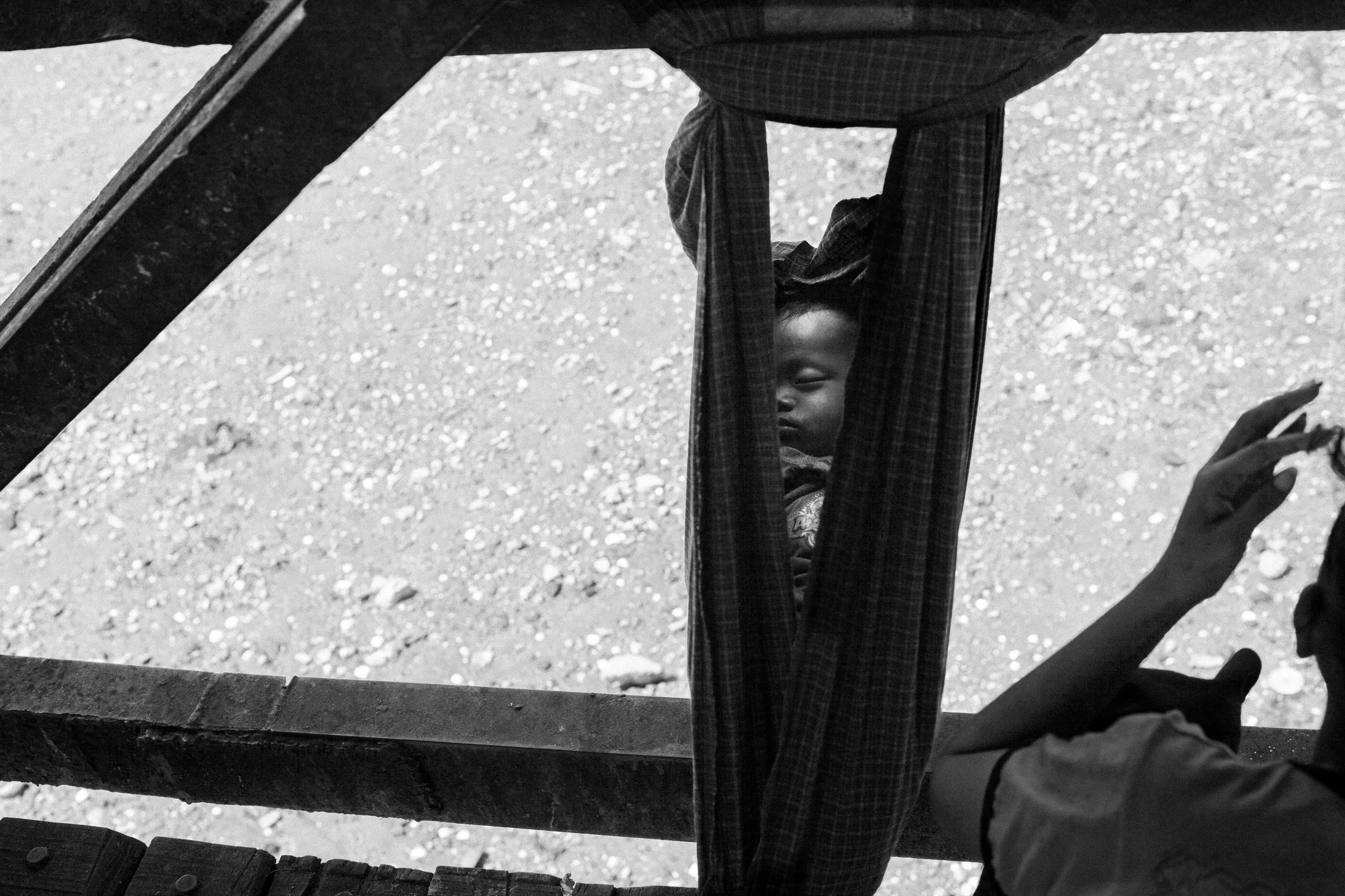 A baby asleep in a cloth sling hung beneath a wooden jetty, a hand reaching toward it