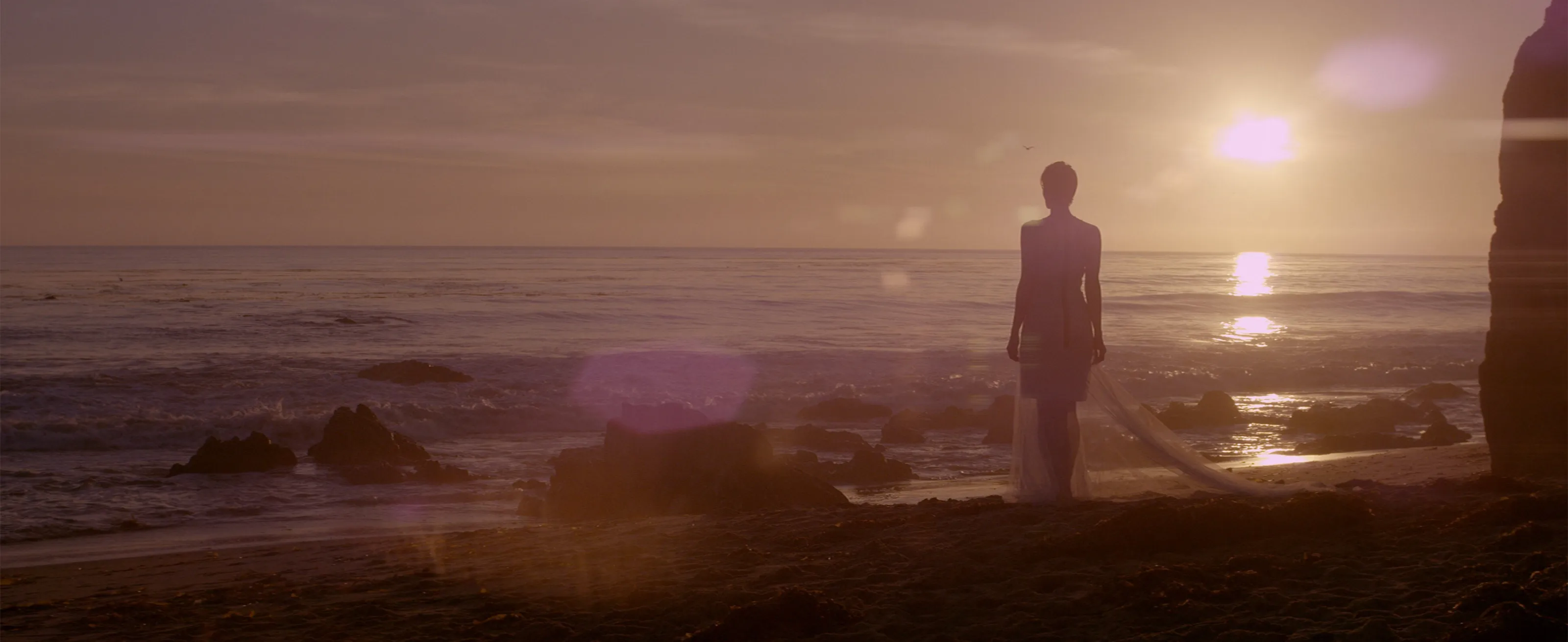 A lone woman on a rocky shoreline at sunset