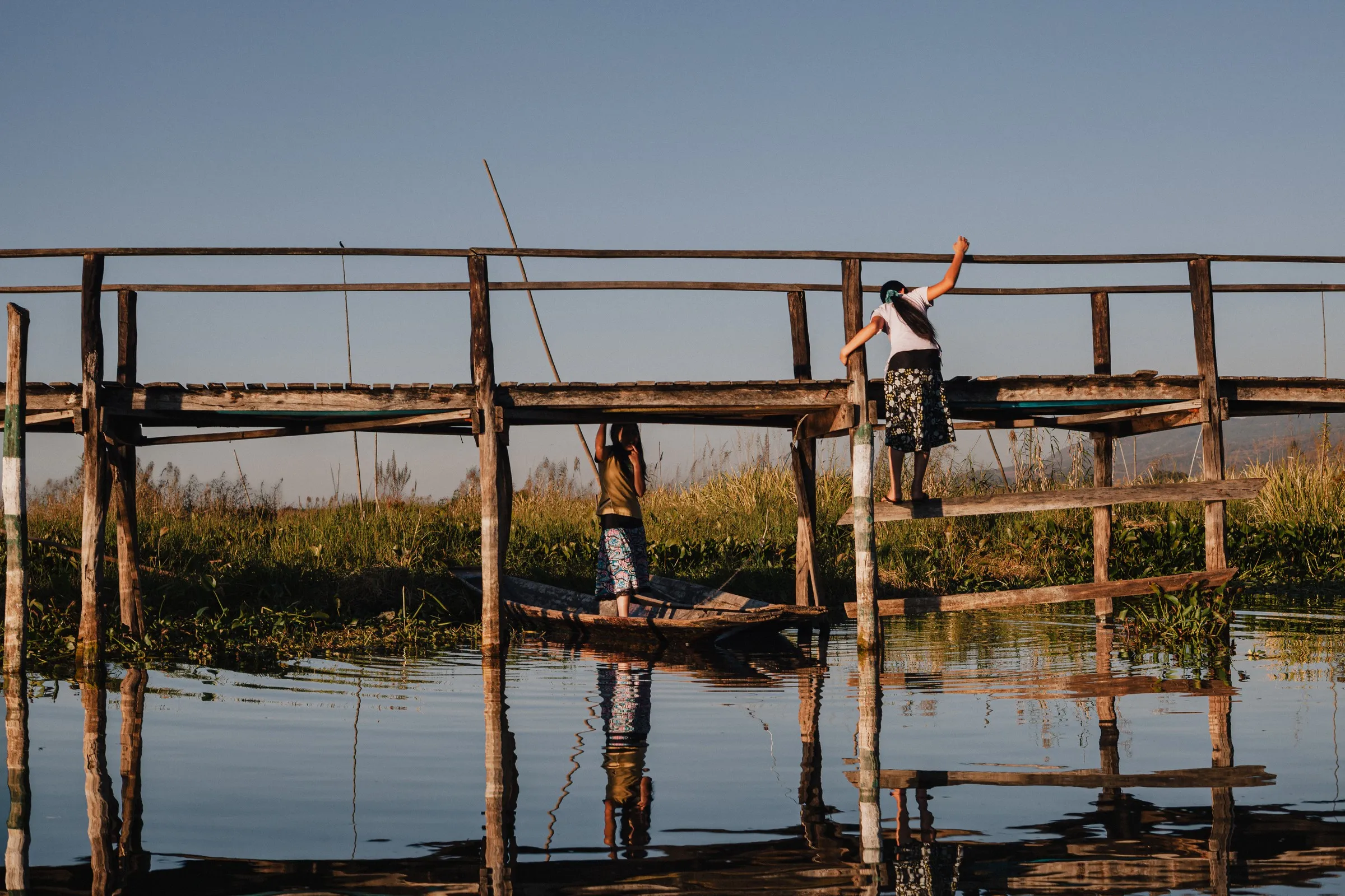 Two girls on a wooden footbridge over the lake, one climbing the rail, their reflections below