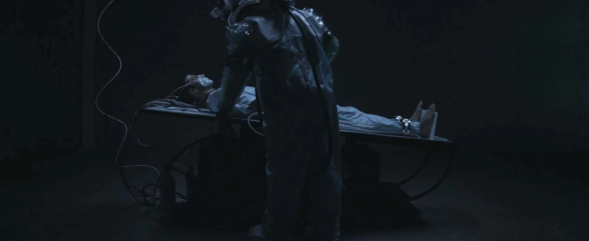In a dark lab, a figure leans over a masked man strapped to a table