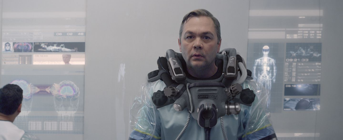 A man in a transparent hazmat suit stands before holographic brain scans