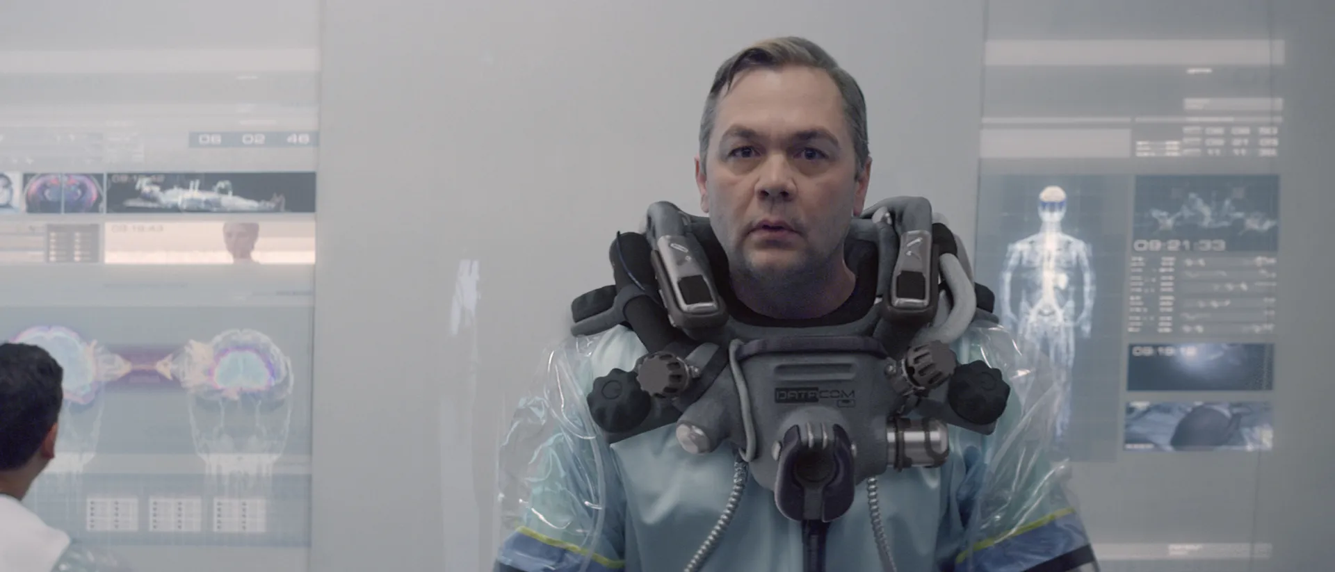A man in a transparent hazmat suit stands before holographic brain scans