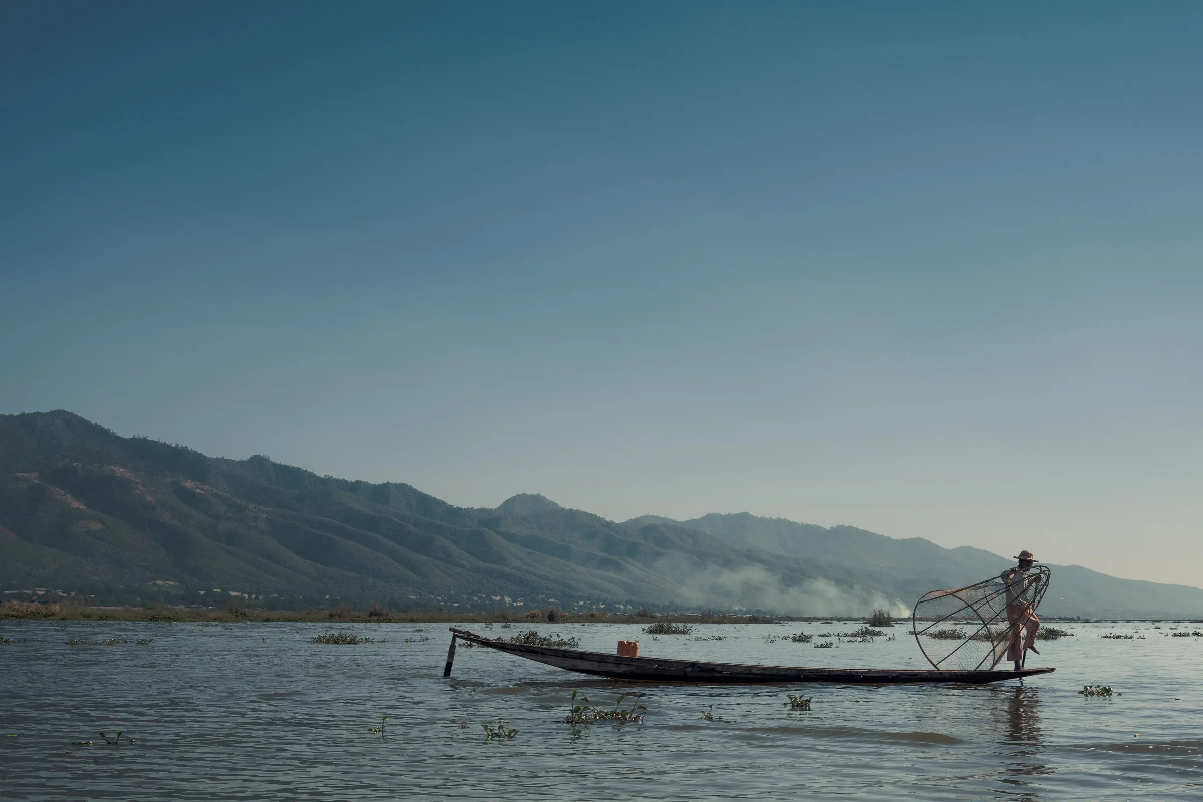 A lone fisherman stands in a long boat on the open lake, hills and haze behind
