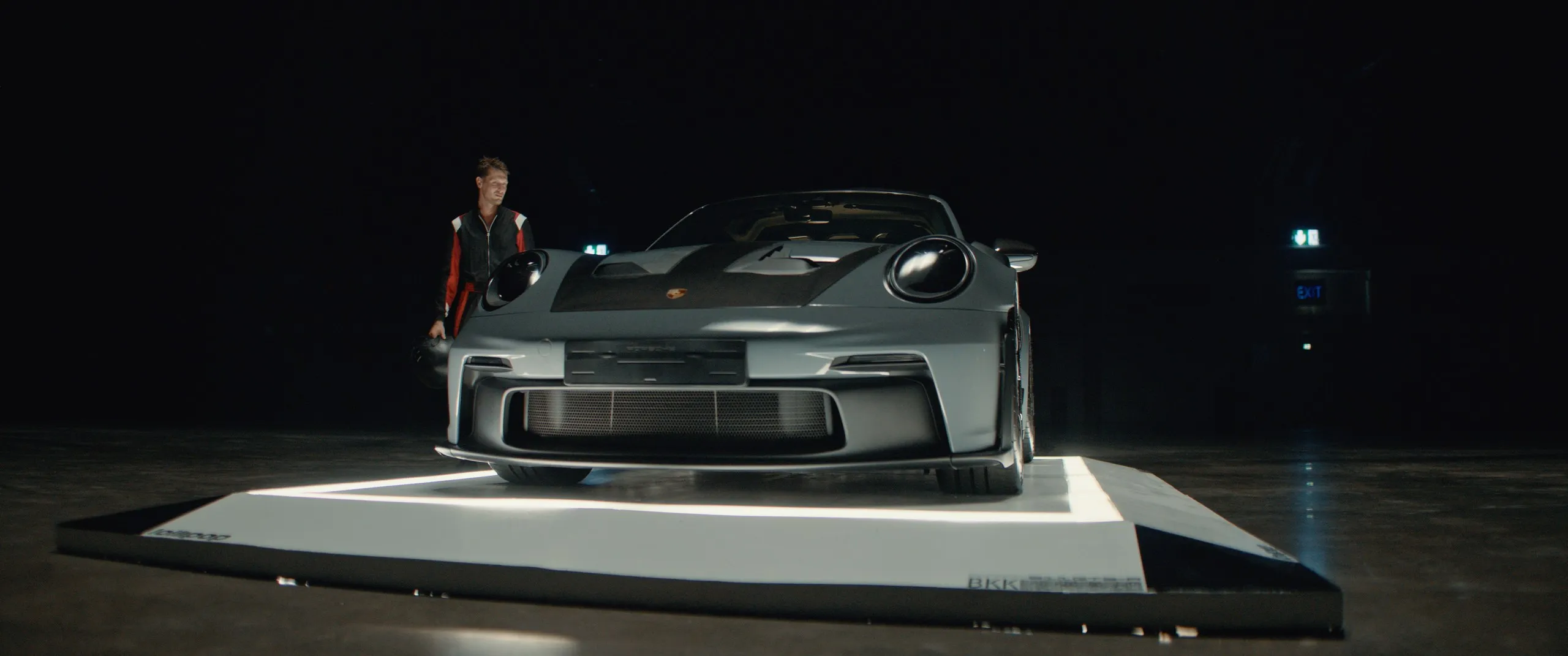 A grey Porsche 911 GT3 RS on a lit platform, a driver beside it
