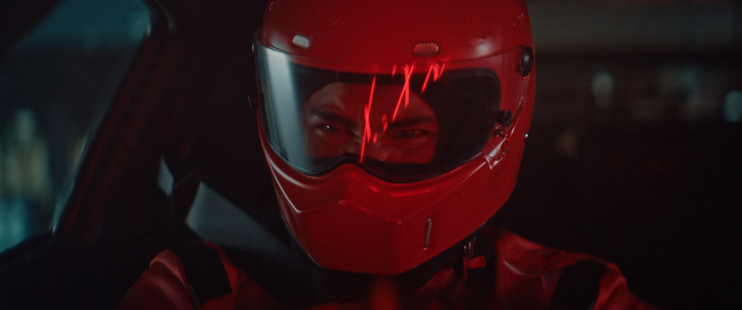 A driver in a red helmet, lit red, behind the wheel